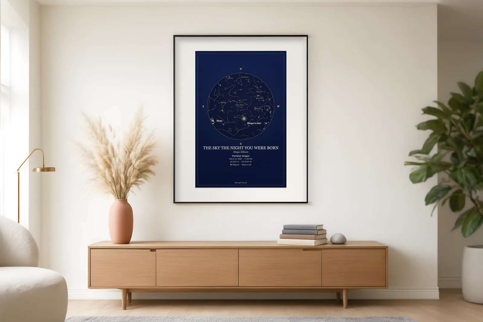 A framed navy star map titled 'The Sky the Night You Were Born' above a low oak sideboard in a bright living room.