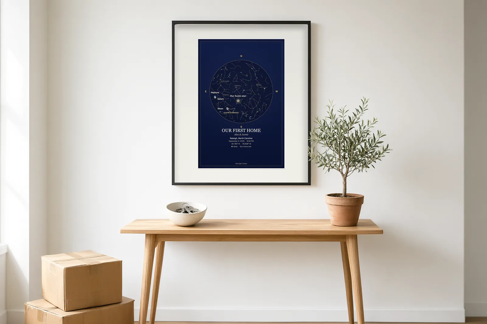 A navy star map titled 'Our First Home' framed in a sunny entryway above a console with a ceramic bowl, a potted olive tree and moving boxes.